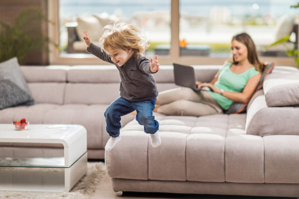 Glad pojke hoppar från soffan hemma. Hans mamma använder en laptop i bakgrunden.