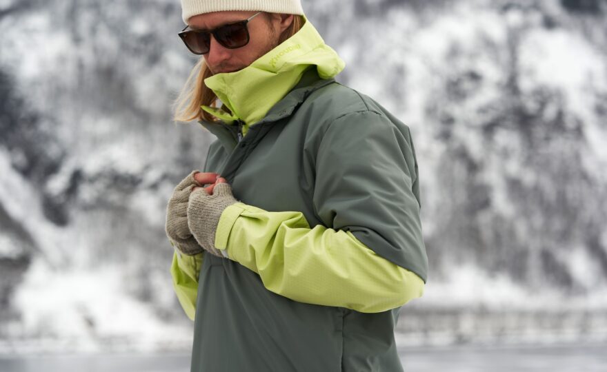 Man med mössa och solglasögon har en grön vinterjacka. I bakgrunden syns ett berg täckt med snö.